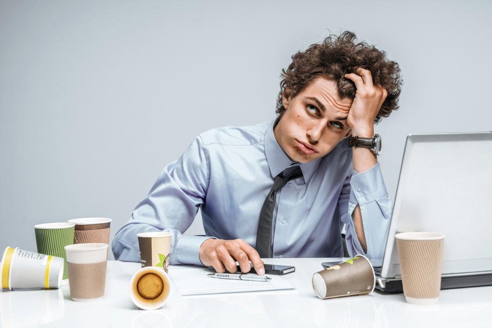 A professionally dressed man grabbing his hair as if stressed and sitting at a laptop computer with cups of coffee everywhere.