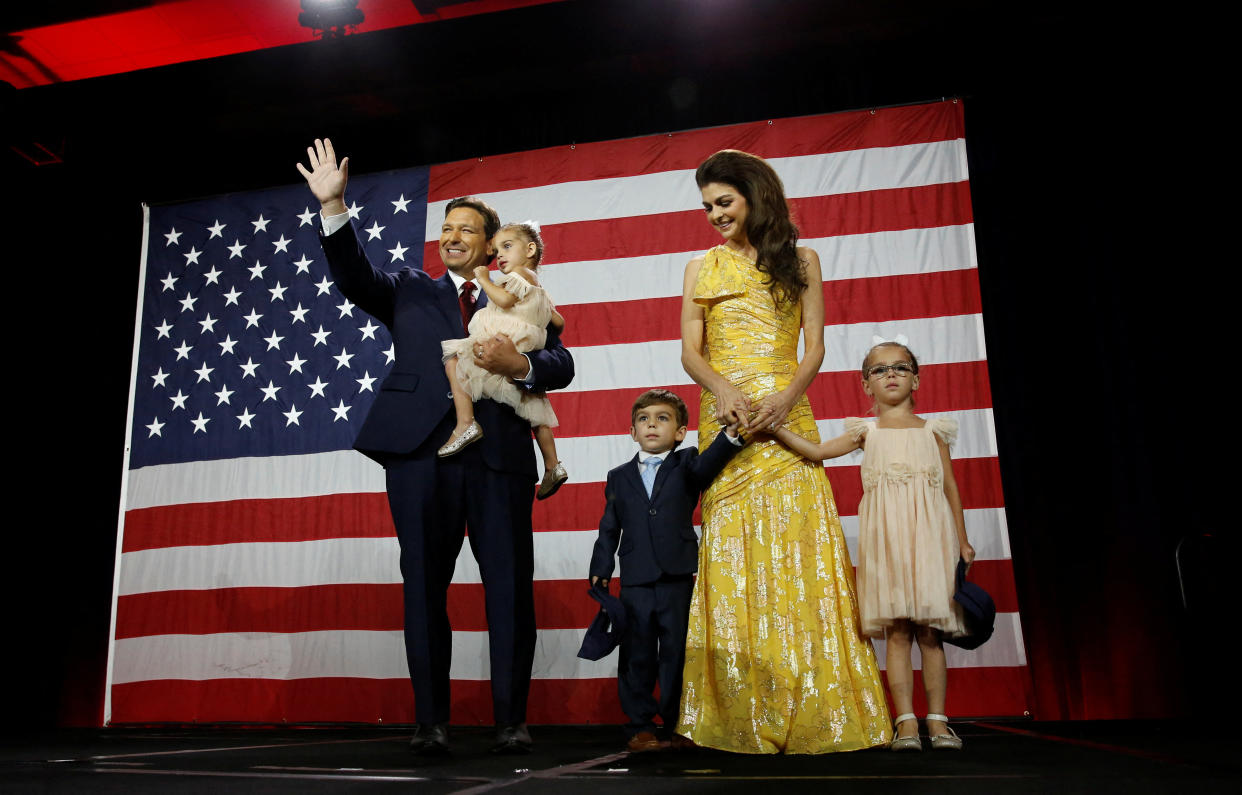 Ron DeSantis waves from stage while holding one child, while his wife stands nearby, holding the hands of two other children.