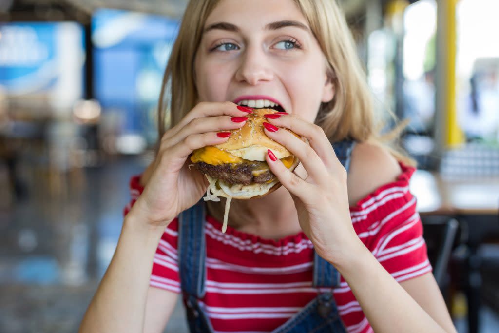A woman eats a burger. She has blonde hair, she's wearing overalls and has red nail polish on her nails. 