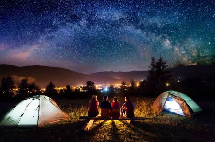 Friends hikers sitting on a bench made of logs and watching fire together beside camp and tents in the night. On the background beautiful starry sky, mountains and luminous town. Rear view