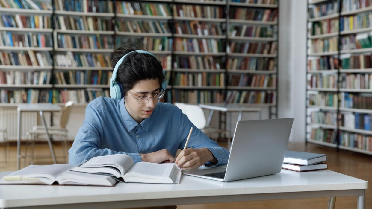  Best laptops for students - Student with long hair is sitting in the library wearing glasses and headphones. Surrounded by books, he's looking at his laptop and writing notes in pencil. 