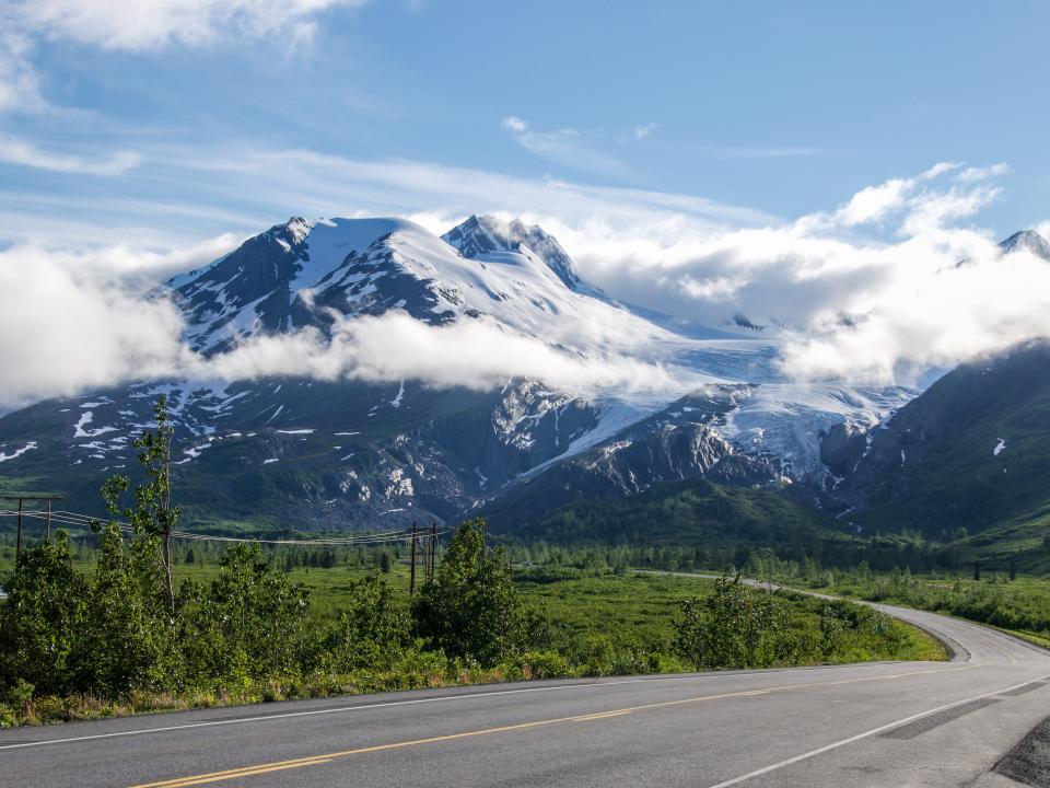 A winding road leading to snow-capped mountains with low clouds. There is green grass on both sides of the road.