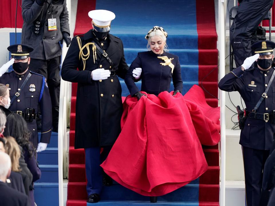 Gaga in a navy jacket top with a giant gold bird broach and a voluminous red skirt being walked down stars by a man in military uniform.