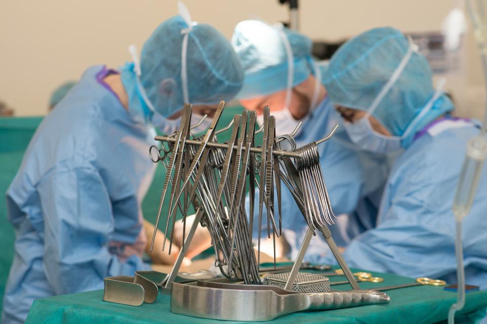 Three health care workers in blue scrubs and face masks are seen behind surgical tools as they focus on something out of view.