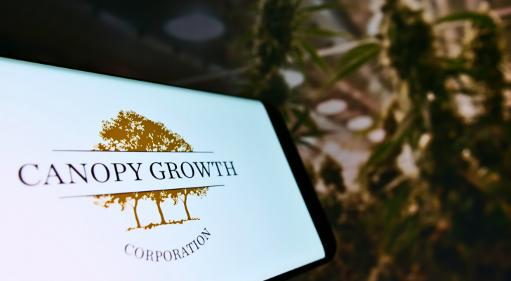 Smartphone with logo of Canadian cannabis company Canopy Growth Corporation on screen in front of web page. Cannabis plants are in the background. CGC stock.