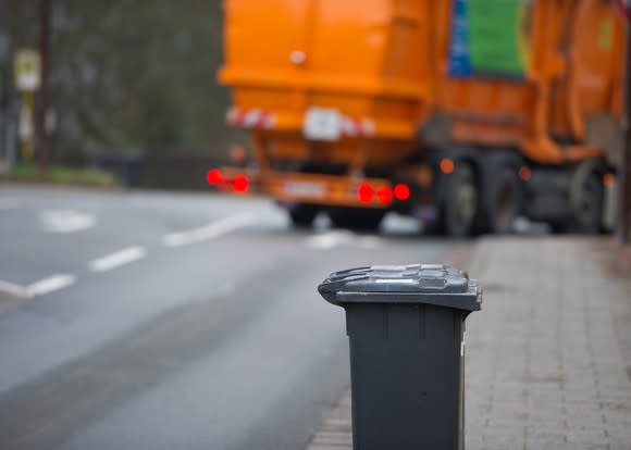 A garbage truck driving away from a garbage can.