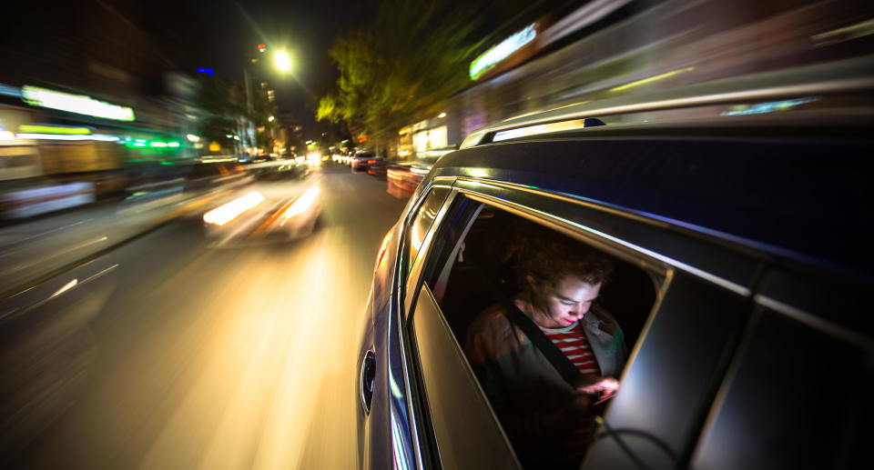 A woman sits in the back of a car, possibly a rideshare, looking at her mobile phone. The lights of New York City streak past. Source: Getty Images