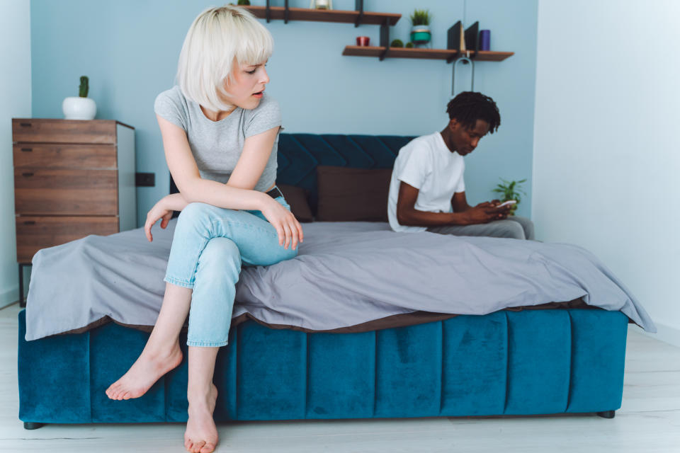 Two people in a bedroom; one seated on bed looking displeased, other in background using phone