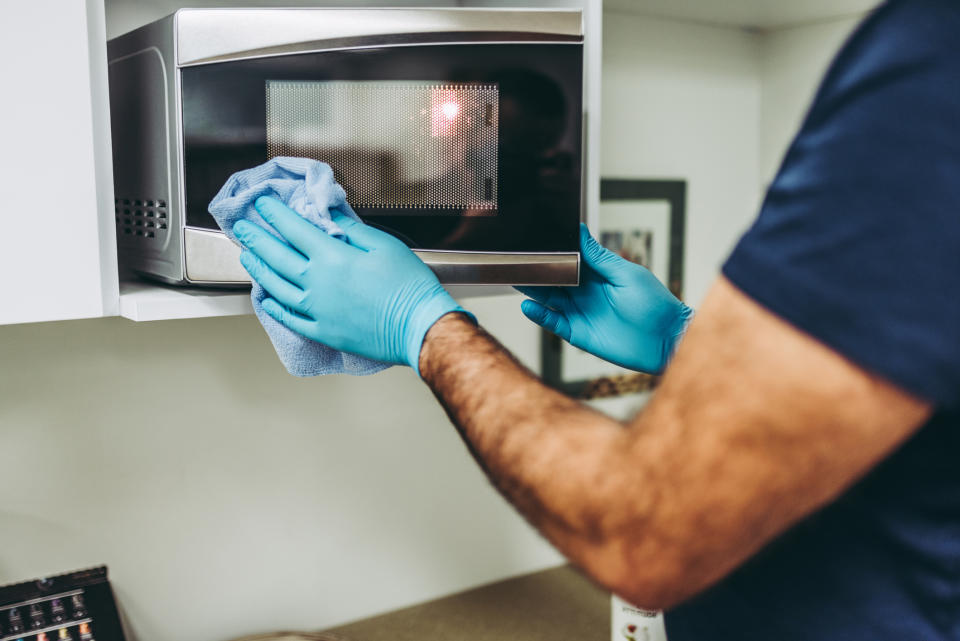 A person wearing blue gloves cleans the exterior of a microwave with a blue cloth