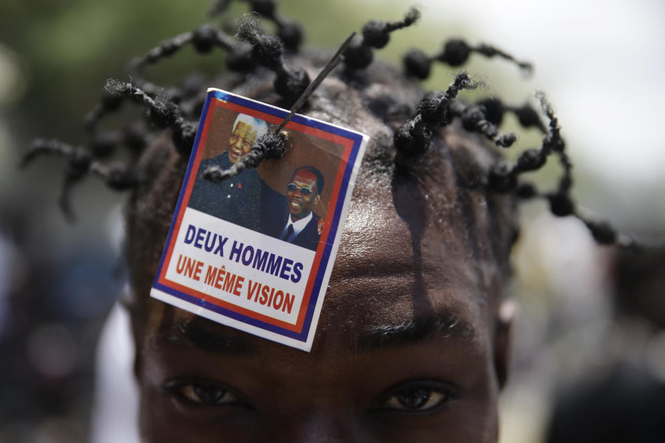 A supporter of former Haitian President Jean-Bertrand Aristide wears a photo of Aristide and late South African President Nelson Mandela as he waits near the airport others for Aristide's expected arrival from Cuba, where he underwent medical treatment in Port-au-Prince, Haiti, Friday, July 16, 2021. Aristide's return adds a potentially volatile element to an already tense situation in a country facing a power vacuum following the July 7 assassination of President Jovenel Moïse. (AP Photo/Joseph Odelyn)