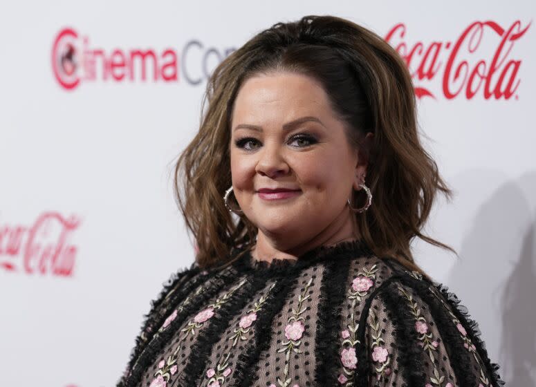 Melissa McCarthy is posing and smiling while wearing a black flowered dress with her hair up.