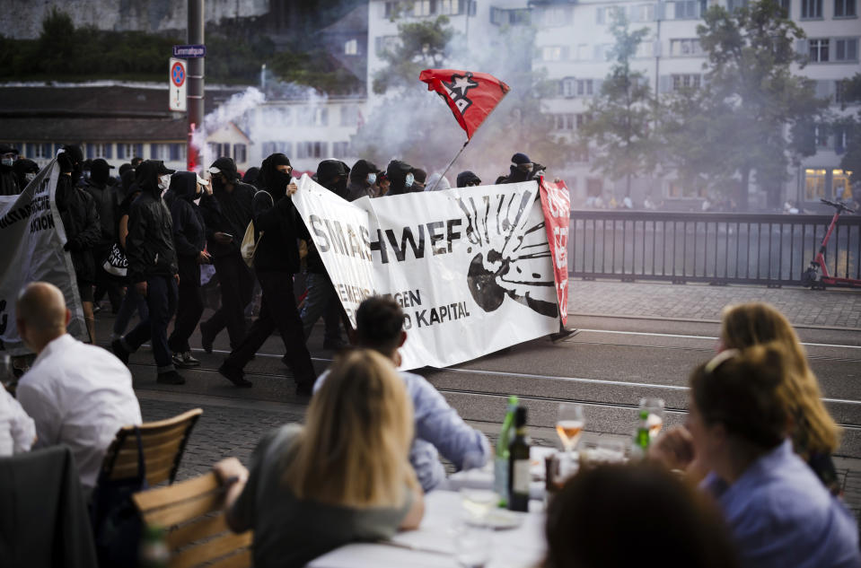 Protesters walk with banners reading "Smash WEF" past people sitting in a restraurant during a demonstration against the World Economic Forum (WEF) in Zurich, Switzerland, Friday, May 20, 2022. The World Economic Forum Annual Meeting will take place from May 22-26, in Davos. (Michael Buholzer/Keystone via AP)