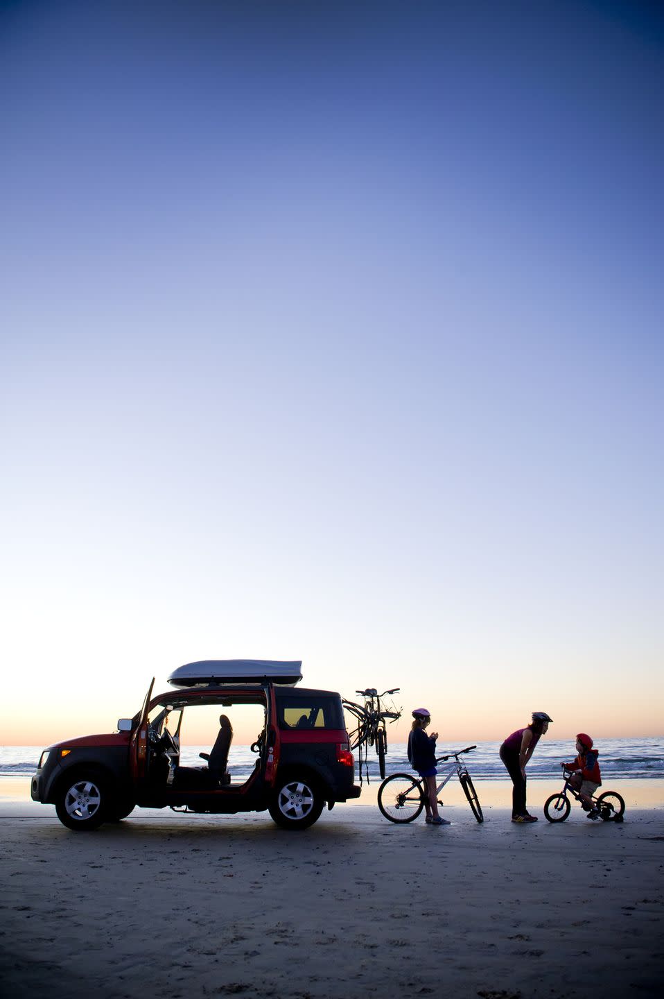 silhouette of mom, dad and child on beach with car and bikes, sunset