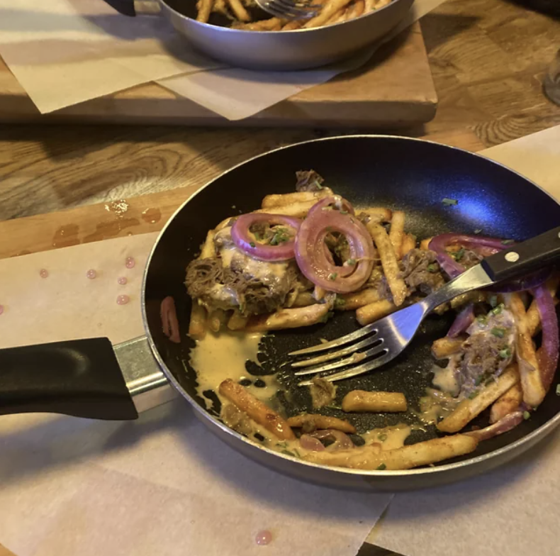 Pan with fries, melted cheese, caramelized onions, and pulled pork. Fork rests inside pan. Background includes another dish and a wooden table