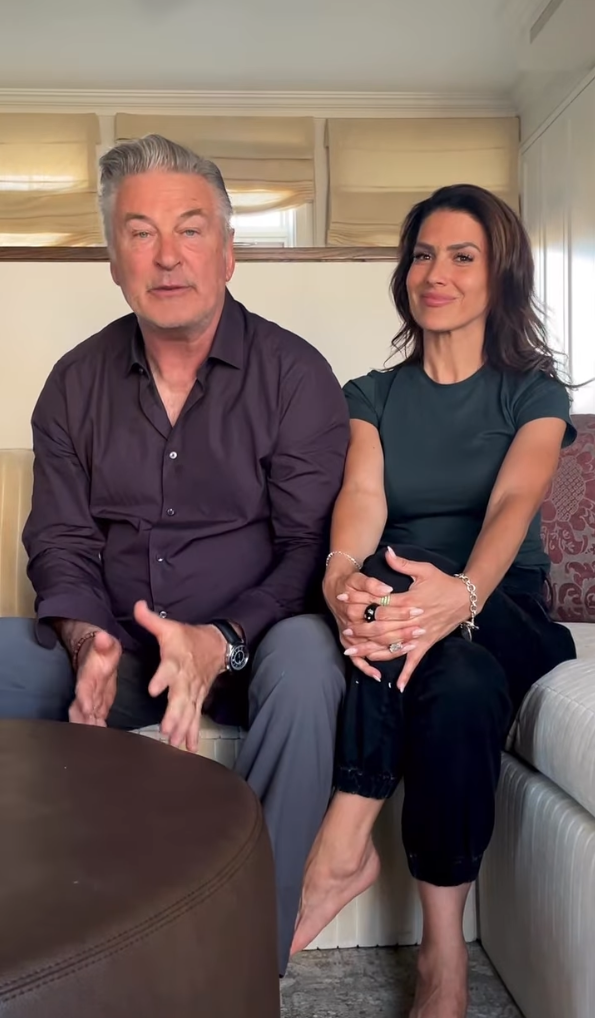 A man and woman, Alec Baldwin and Hilaria Baldwin, sitting on a couch. Alec is wearing a button-up shirt, Hilaria is wearing a casual top and pants