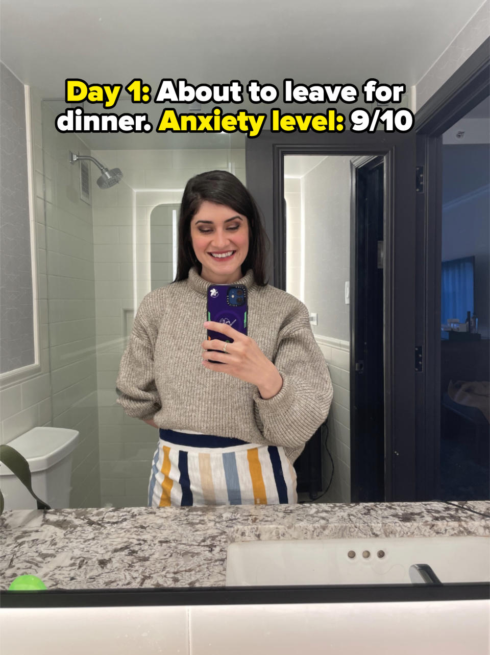 Woman smiling in a bathroom mirror, wearing a gray knit sweater and striped skirt