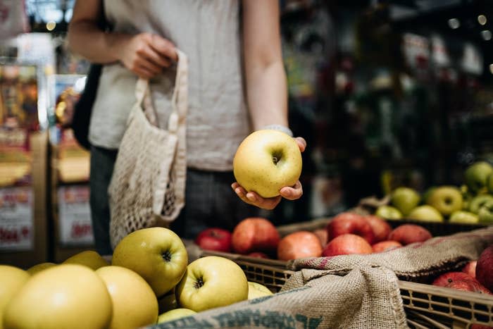 Person holding a yellow apple, shopping at a market stand with various apples