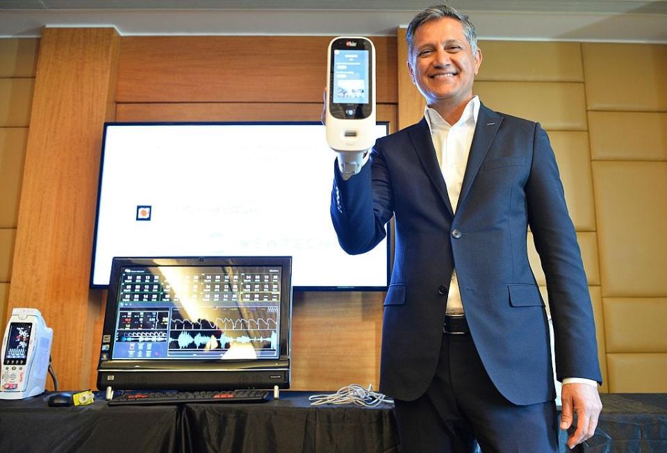 Joe Kiani holding up a medical device in front of a series of screens showing data