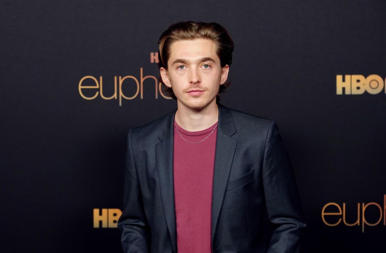 austin abrams, a young man stands looking at the camera, brown hair slicked back, wearing red tshirt and black jacket