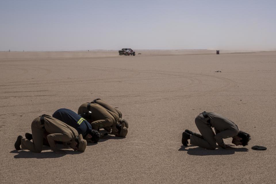 In this Sunday, Jan. 12, 2020 photo, Saudi Arabia security forces pray while a care races during stage seven of the Dakar Rally between Riyadh and Wadi Al Dawasir, Saudi Arabia. Formerly known as the Paris-Dakar Rally, the race was created by Thierry Sabine after he got lost in the Libyan desert in 1977. Until 2008, the rallies raced across Africa, but threats in Mauritania led organizers to cancel that year's event and move it to South America. It has now shifted to Saudi Arabia. The race started on Jan. 5 with 560 drivers and co-drivers, some on motorbikes, others in cars or in trucks. Only 41 are taking part in the Original category. (AP Photo/Bernat Armangue)