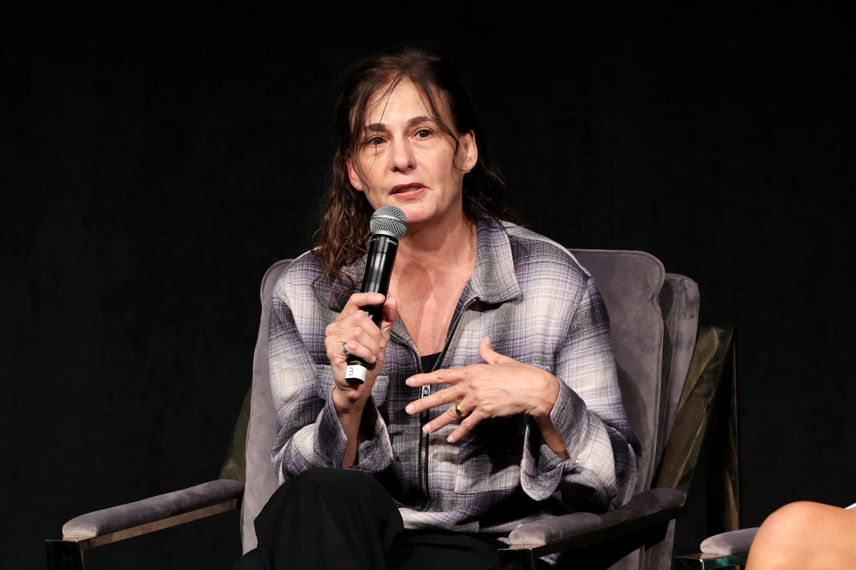 Madeleine Gavin, seated in an armchair against a black background, holds a microphone to her mouth.