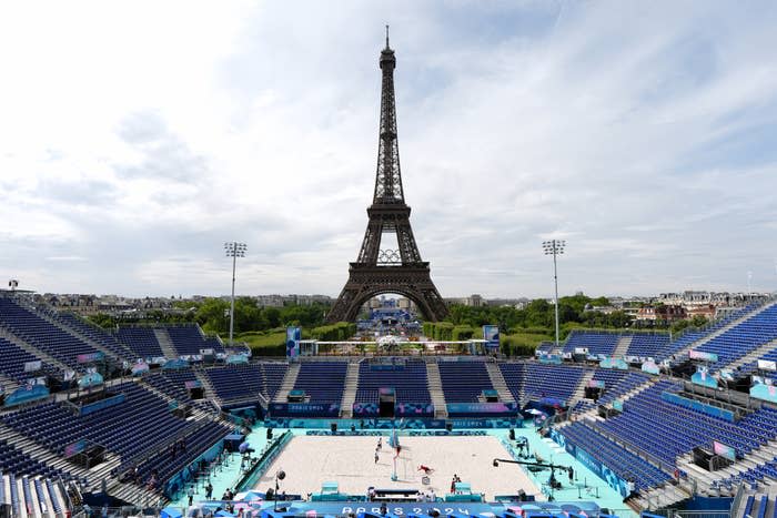 Eiffel Tower in the background of a Paris 2024 Olympic Games volleyball stadium setup, with players practicing on the sand court. Stadium seats are mostly empty