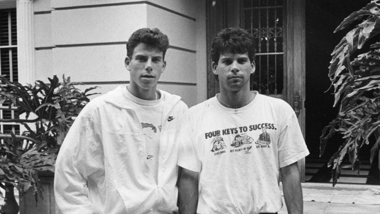 erik and lyle menendez stand outside a home and stare at the camera, the front door is open, erik wears a jacket over a tshirt with shorts, lyle wears a tshirt with pants