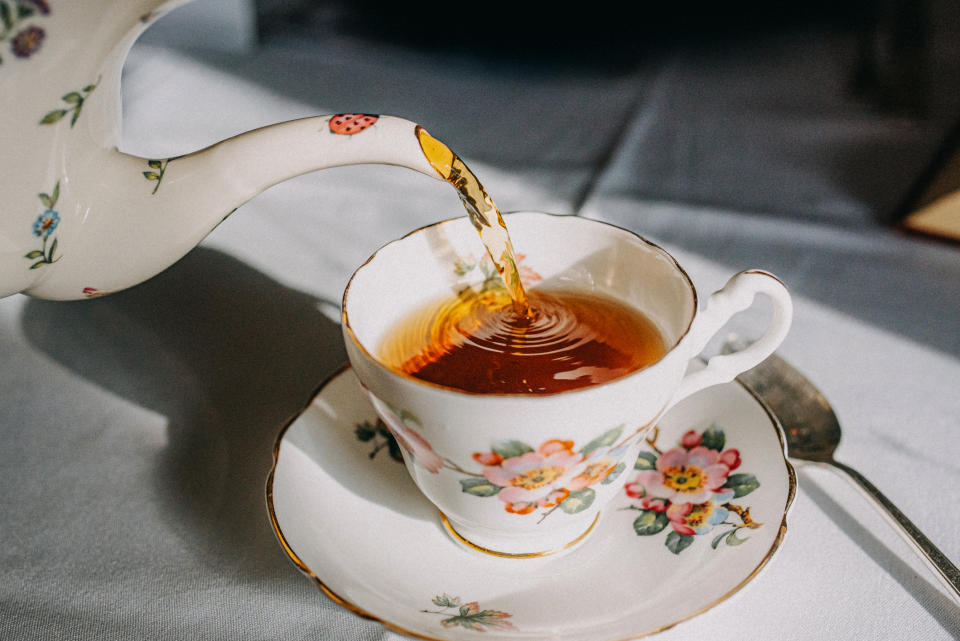 A porcelain teapot with delicate floral patterns pours tea into a matching teacup on a saucer. A spoon lies next to the saucer