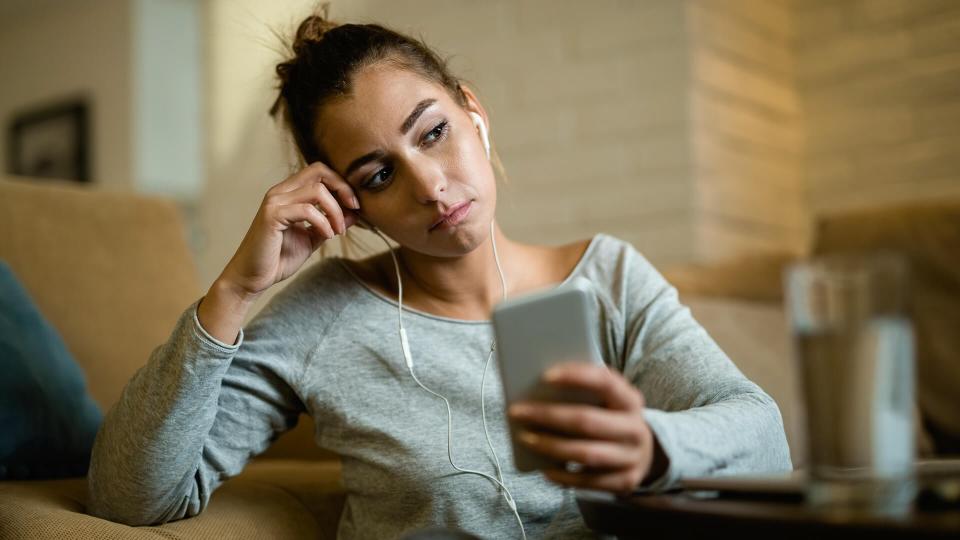 Young pensive woman feeling sad while using smart phone in the evening at home.
