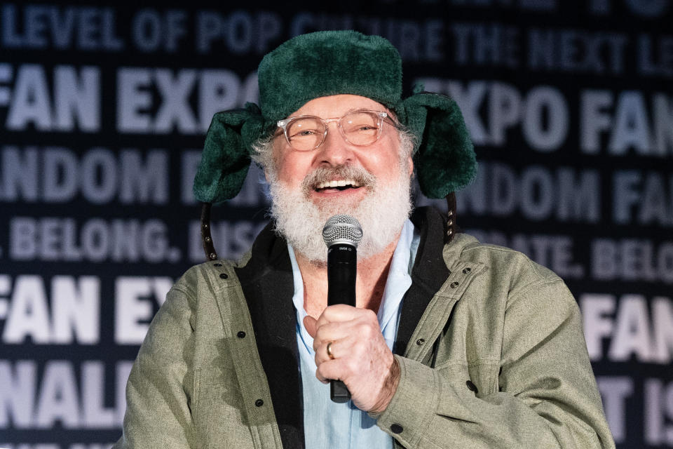 Person on stage speaking into a microphone, wearing a whimsical hat with ear flaps