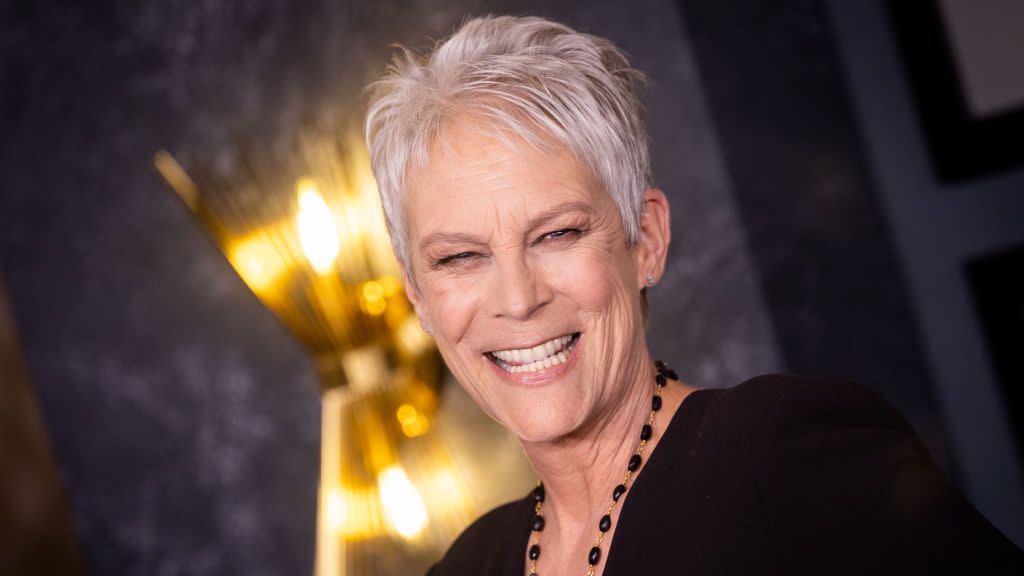 jamie lee curtis smiles and looks toward the camera, she wears a black outfit with a beaded matching necklace, a gold statute is in the background