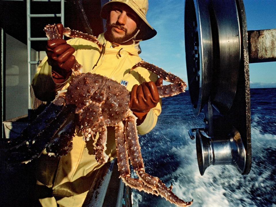 fisherman in yellow waterproof jacket holds up giant king crab aboard fishing boat