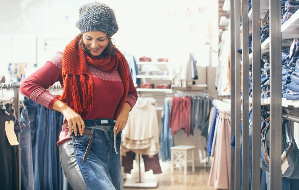 Closeup of smiling adult woman trying on new pair of jeans in shopping mall. She has put the pants over her legs to se if they fit. She's wearing red cardigan and scarf and gray hair. There are many different jeans around her.