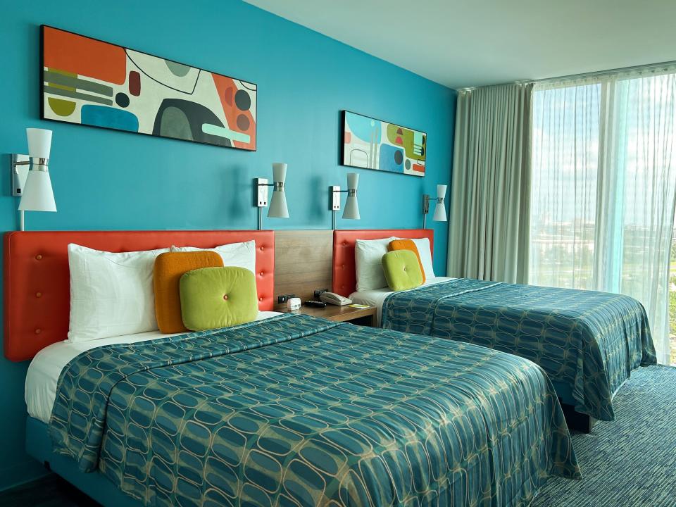 A hotel room with blue walls, retro paintings, and two full-sized beds. The beds have three pillows - one white, one orange, and one green. The comforters are blue with a pattern. The headboard is red. There is a floor-to-ceiling window with curtains on the right.