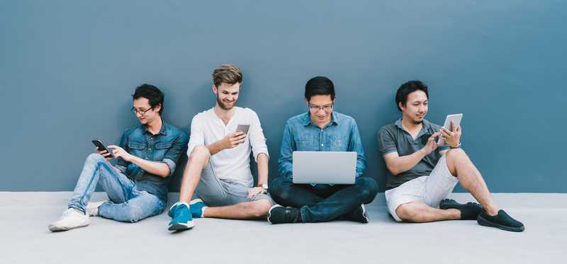 .Four young adults sitting against a wall using various smart devices.