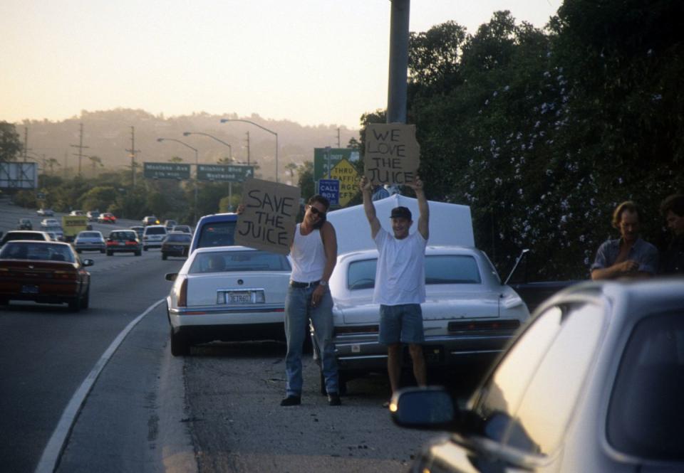 two men stand on a highway shoulder and hold cardboard signs with messages, cars drive past