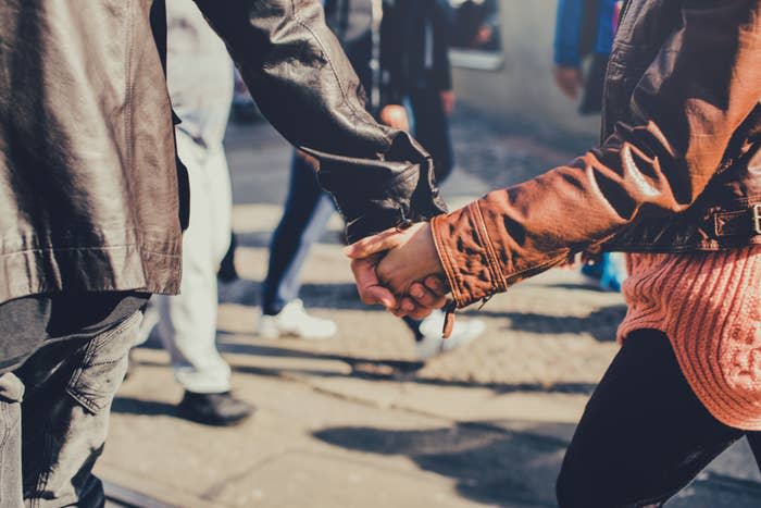 A couple is holding hands while walking in a crowded area. They are wearing casual clothes, including leather jackets. Individuals in the background are blurred