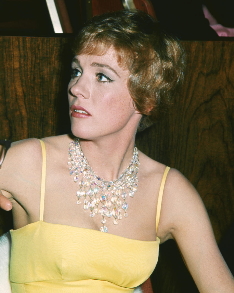 A woman in a sleeveless dress wears an ornate, multistrand diamond necklace and looks to the side. She has short hair and is seated indoors