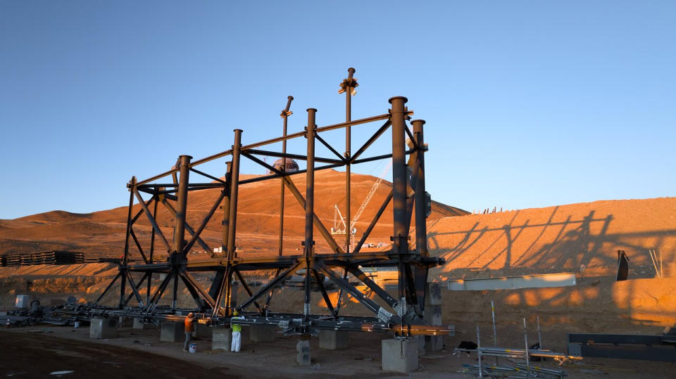 On a hillside in a mountainous desert, large metal beams construct the framework for a large structure.