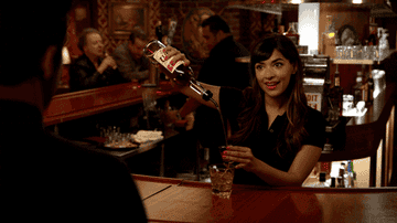 Woman behind bar pouring drink, smiling at someone off-screen, in a scene from TV show