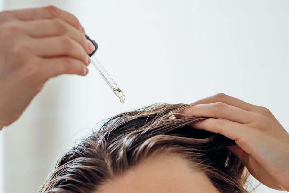 A person applies hair oil using a dropper onto their scalp. Their other hand is parting their hair to aid the application