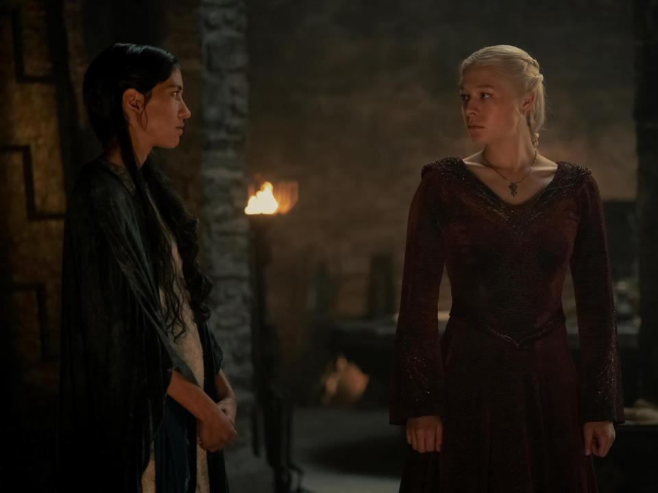 sonoya mizuno and emma d'arcy as mysaria and rhaenyra targaryen. mysaria is dressed in common clothing, standing to the side, while rhaenyra is wearing a deep red dress and looking towards her