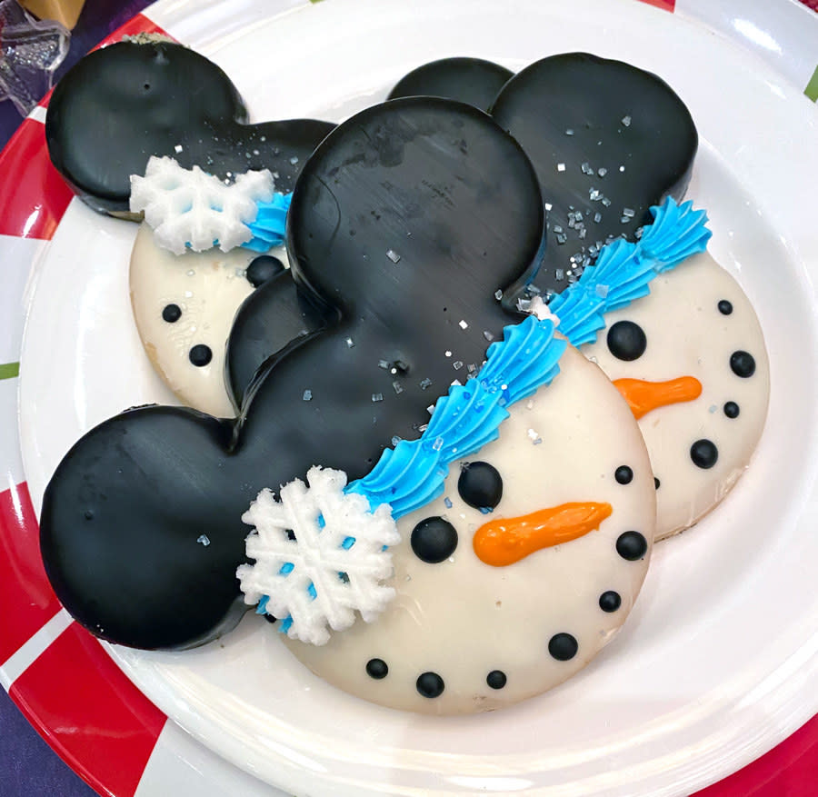 A mickey shaped cookie with the face decorated like a snowman with a carrot nose and coal shaped eyes and mouth and a snowflake