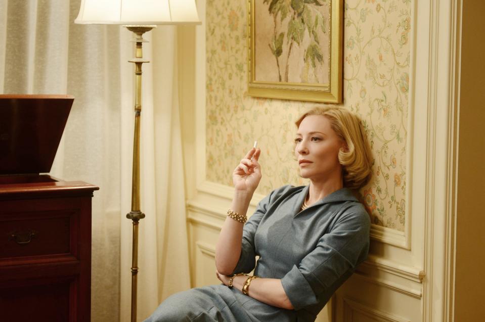 Cate Blanchett in a vintage-style dress, leaning against a wall, holding a cigarette indoors