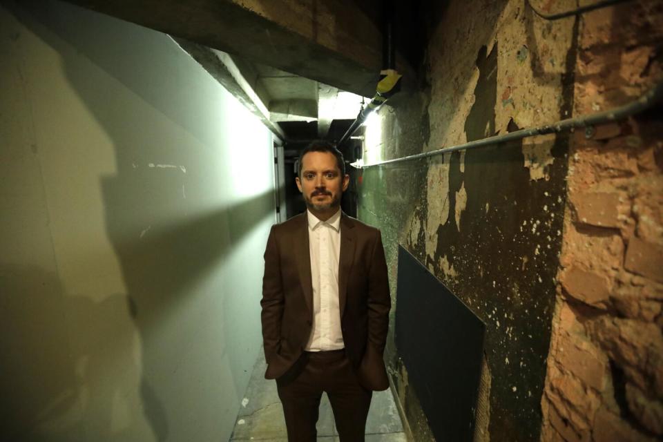 Elijah Wood stands in a hallway with his hands in his pockets.