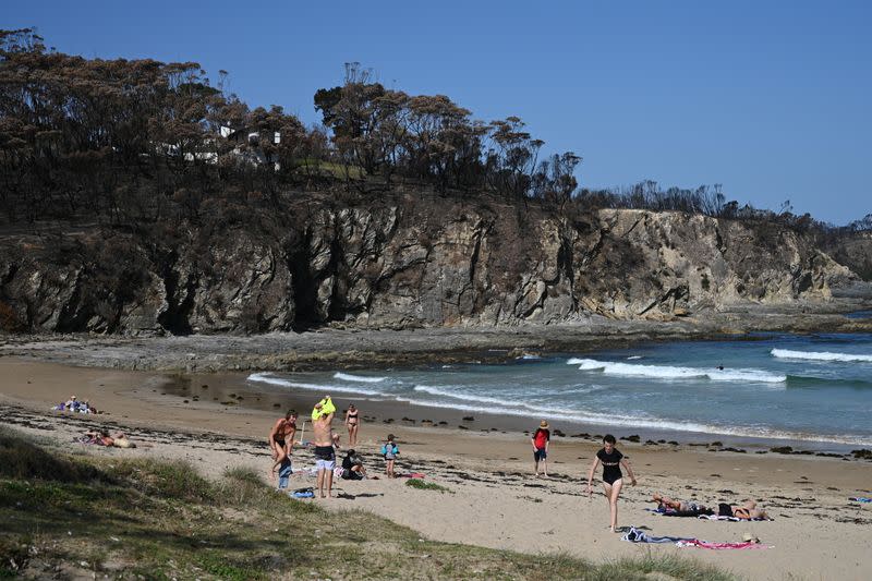 A charred hillside, burnt during the recent bushfires, is pictured behind beachgoers in Malua Bay