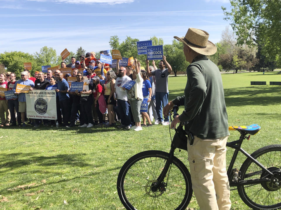 In this Saturday, June 1, 2019 photo, a passing bicyclist pauses to watch supporters of Denver Mayor Michael Hancock as they pause for a picture during a rally for the incumbent in Denver. Hancock, who is seeking his third, four-year term as mayor, is facing Jamie Giellis in a runoff Tuesday. (AP Photo/James Anderson)