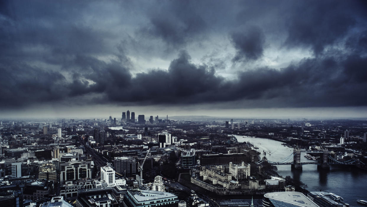 Aerial view of London looking down the River Thames towards Canary Wharf and the Docklands. In the foreground is the Tower of London and Tower Bridge. A dark and menacing sky is overhead.