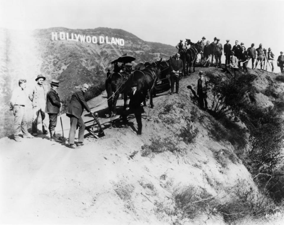 A historical photo of people gathered around a camera and equipment on a hillside with the "Hollywoodland" sign in the background