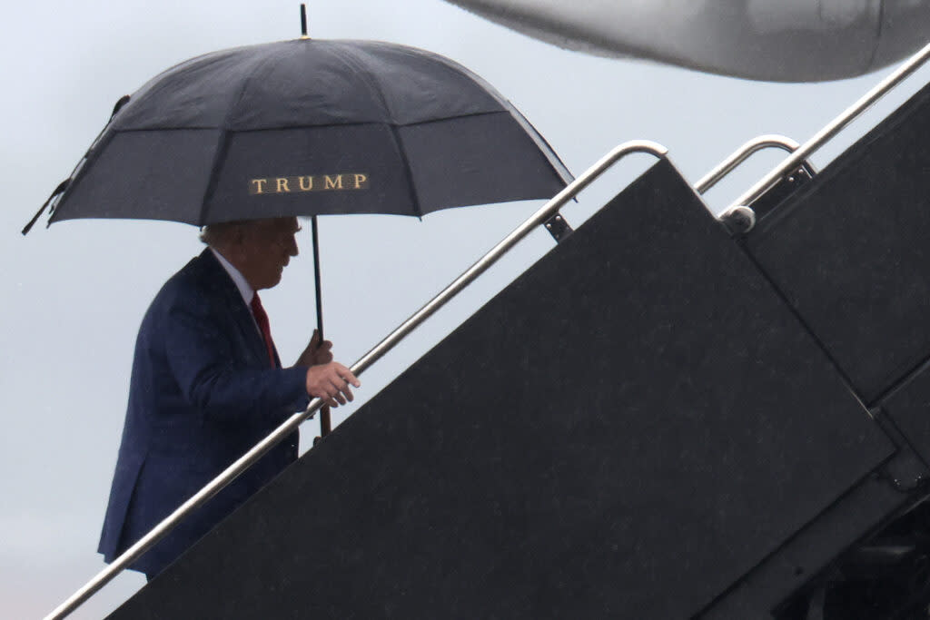 Donald Trump, holding a large black umbrella with a gold "TRUMP" written on the side, walks up the steps to an airplane.